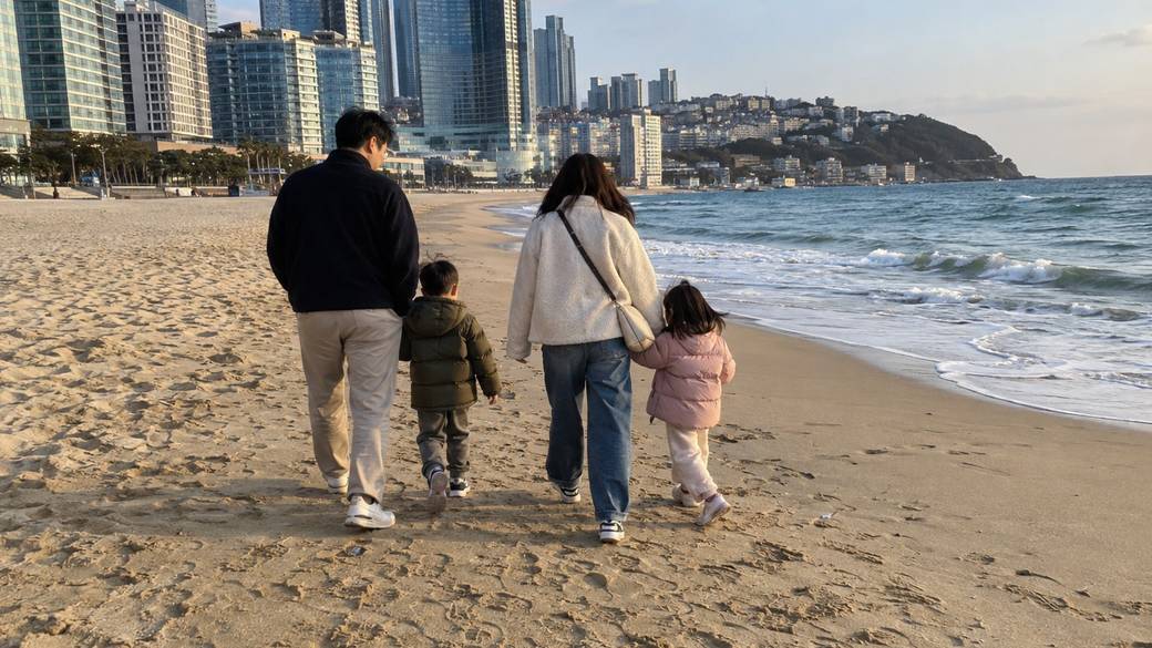 A family with two young children walking along the sand at Haeundae Beach in Busan in the late afternoon, hotels behind them