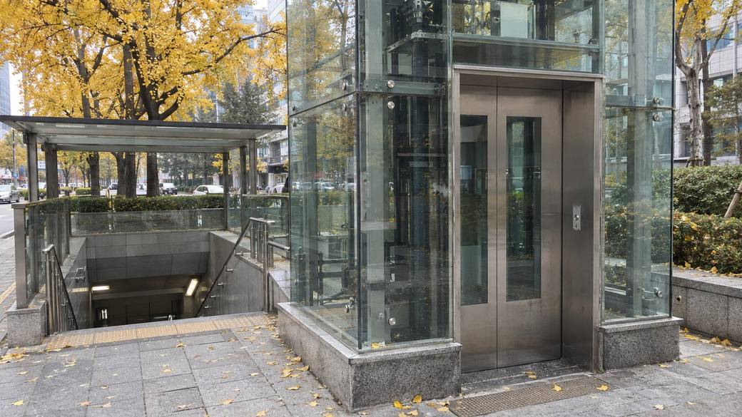 A glass street-level subway elevator beside a station entrance on a wide Seoul pavement, autumn ginkgo trees behind it